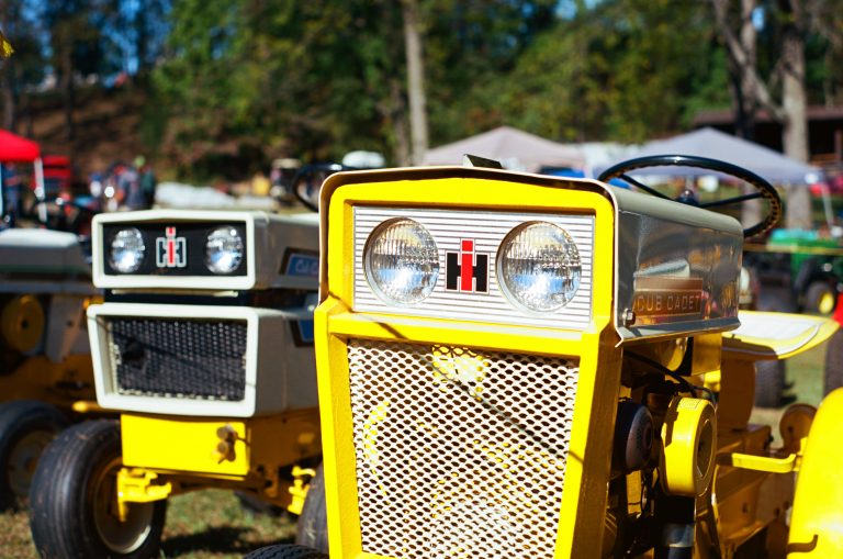Two vintage yellow and white International Harvester Cub Cadet tractors are parked outdoors on grass, with trees and tents visible in the blurred background. The focus is on the front grills and headlights. | Photo by Millworks Film Photography & Prints