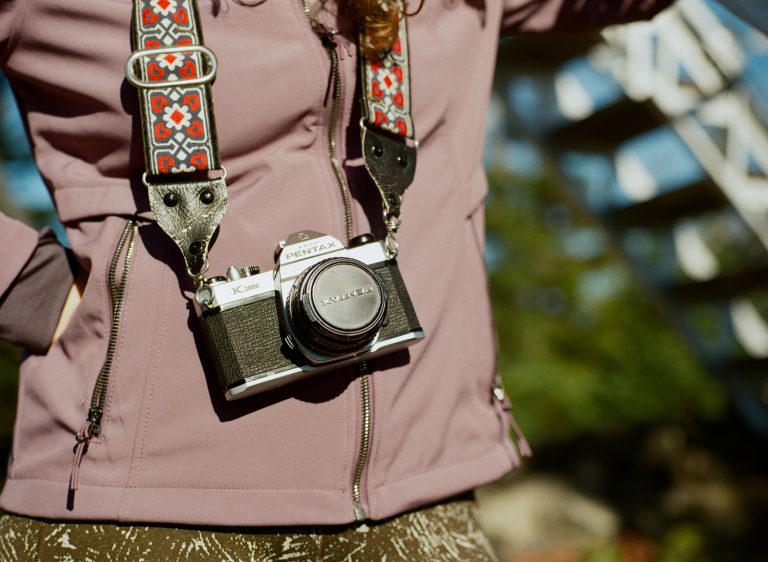 A person wearing a purple jacket and patterned camera strap stands outdoors with a vintage Pentax camera hanging from their neck, ready to capture the beauty of nature and landscapes. The background is softly blurred with natural light and greenery. | Photo by Millworks Film Photography & Prints