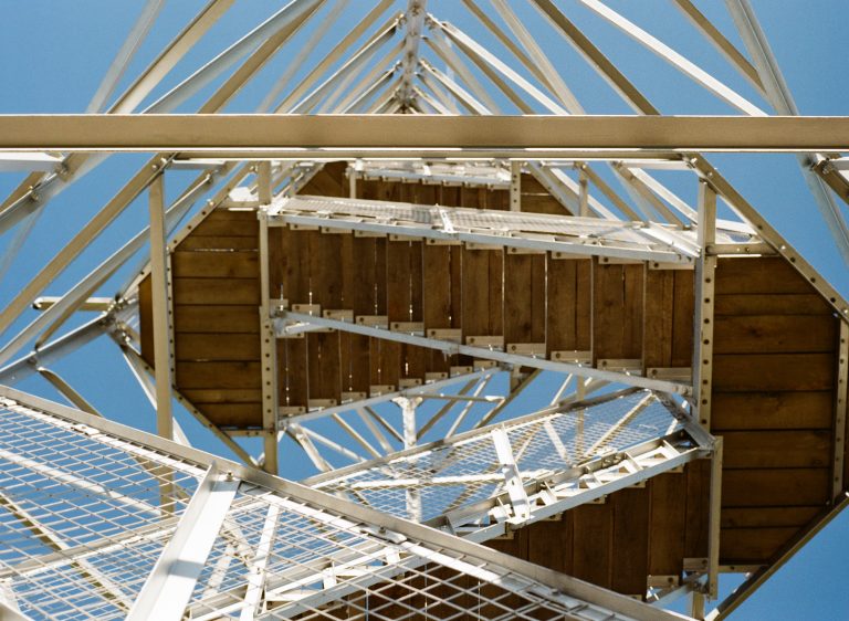 A view from beneath a metal observation tower, set amidst nature, showing wooden steps, metal railings, and beams against a clear blue sky. The geometric structure creates an intricate, angular pattern in scenic landscapes. | Photo by Millworks Film Photography & Prints