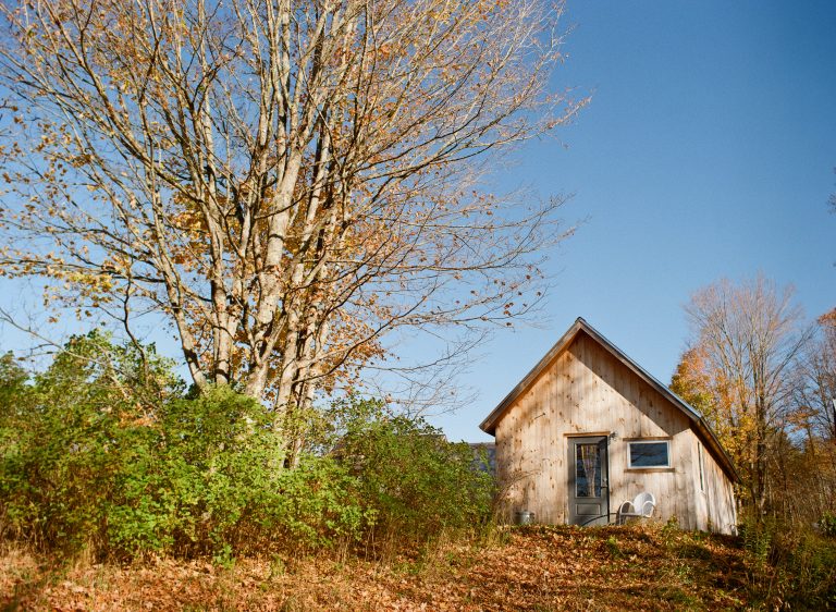 A small wooden cabin sits on a grassy hill next to a leafless tree, surrounded by autumn landscapes and vibrant nature under a clear blue sky. Two white chairs are placed near the cabin’s entrance. | Photo by Millworks Film Photography & Prints