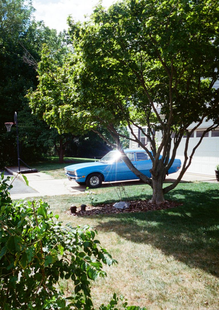 A bright blue vintage car is parked in a driveway beside a white house, partially shaded by a leafy tree. Sunlight reflects off the car’s window. Green grass and shrubs surround the scene on a clear day. | Photo by Millworks Film Photography & Prints