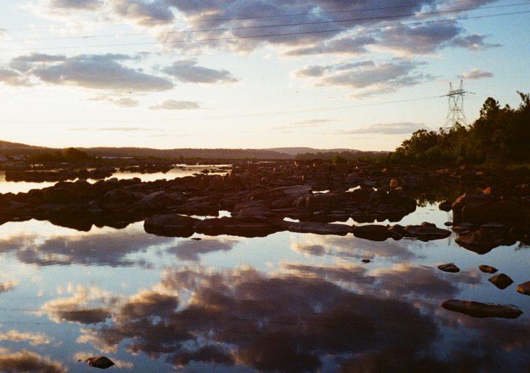 A rocky river reflects clouds and a colorful sunset sky, with distant hills, power lines, a metal tower, and trees on the right, creating a peaceful, natural landscape scene. | Photo by Millworks Film Photography & Prints