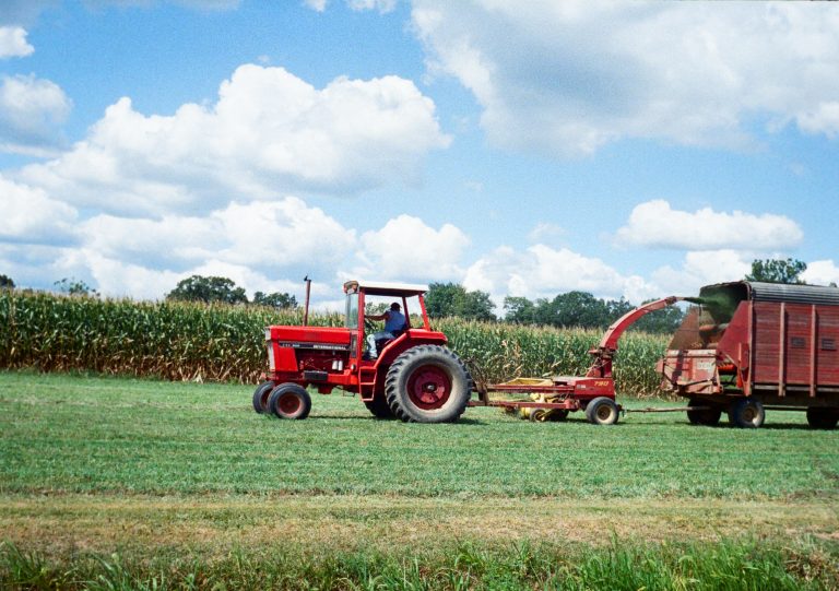 A red tractor pulls a farming machine and a trailer across a green field, with tall corn growing in the background under a blue sky with scattered clouds. | Photo by Millworks Film Photography & Prints